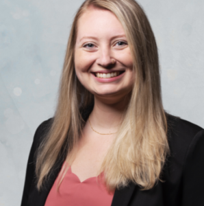 A smiling woman with long blonde hair wears a black blazer over a pink top, standing in front of a light grey background.