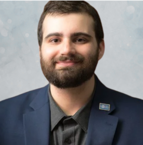 A man with brown hair and a beard smiles, wearing a navy blazer and dark shirt. He has a pin on his lapel. The background is light and softly blurred.
