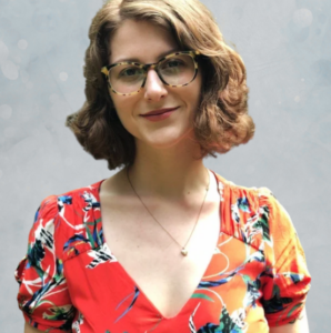 A woman with short wavy brown hair and glasses smiles while wearing a red floral dress and a necklace, standing in front of a light gray background.