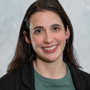 A woman with long brown hair, wearing a green shirt, black blazer, small hoop earrings, and a nose ring, smiles at the camera against a light, softly patterned background.